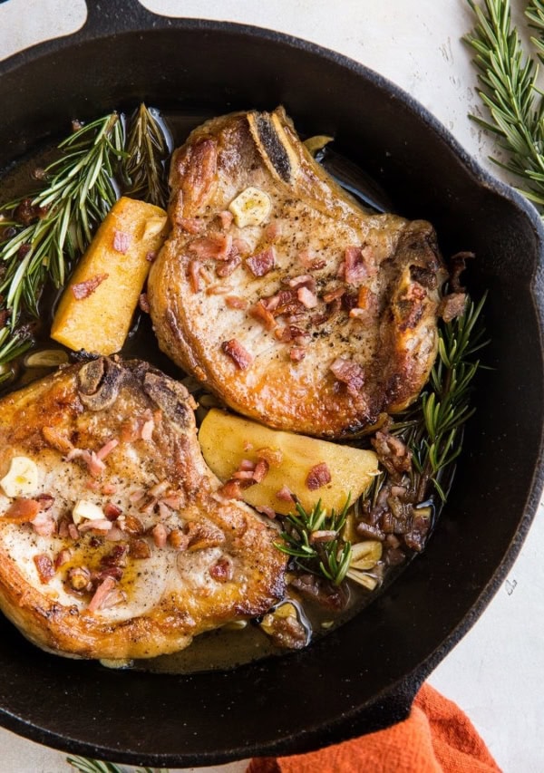 Roasted Pork Chops with cider-spiced apples, bacon, roasted garlic, and rosemary in a cast iron skillet on top of a white backdrop with a red napkin