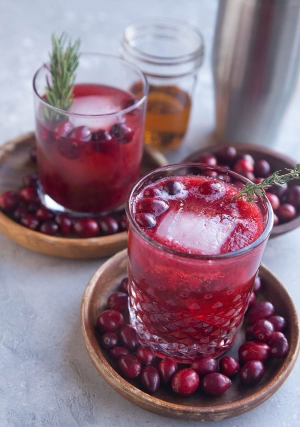 Two cranberry cocktails on two wooden plates with fresh cranberries all around and a cocktail shaker in the background.