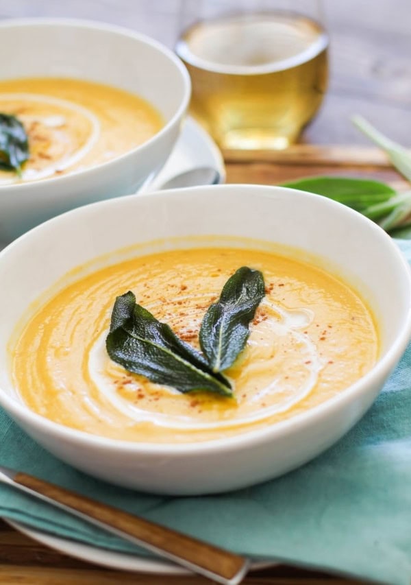 Two white bowls with butternut squash soup on a wooden cutting board with wood inlay spoons and white wine in the background.