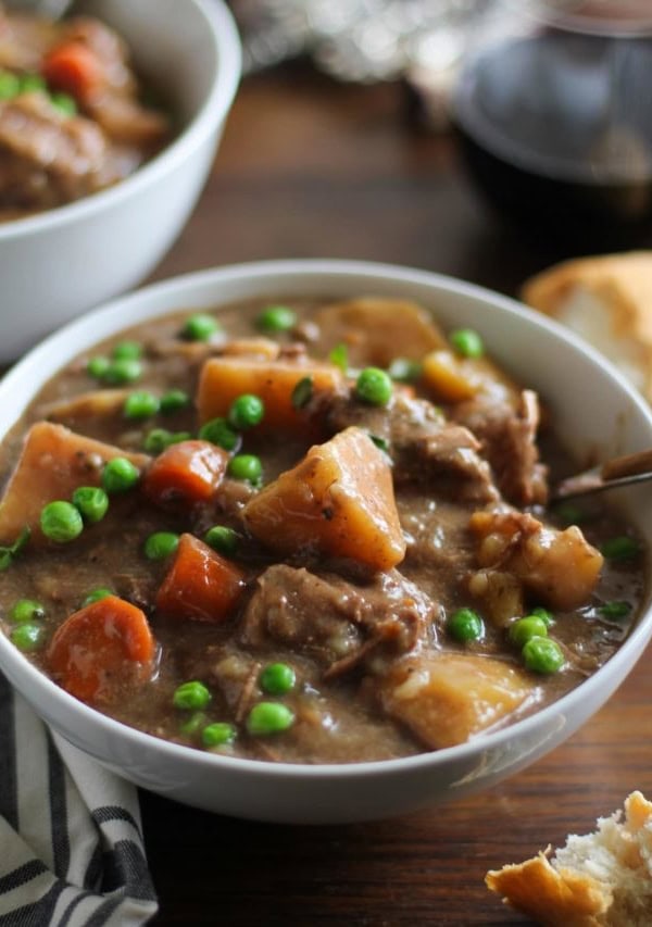 two white bowls of hearty beef stew on a rustic wood background with bread and wine to the side.