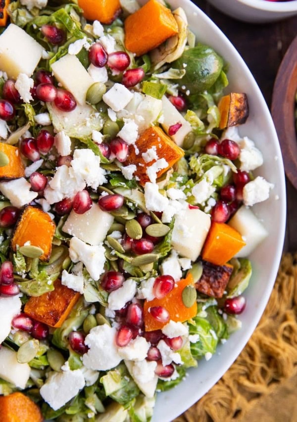 Close up top down photo of white bowl of Shaved Brussel Sprout Salad with Roasted Butternut Squash, Pomegranate Seeds, Pumpkin Seeds, Feta, and Citrusy Maple Cinnamon Dressing