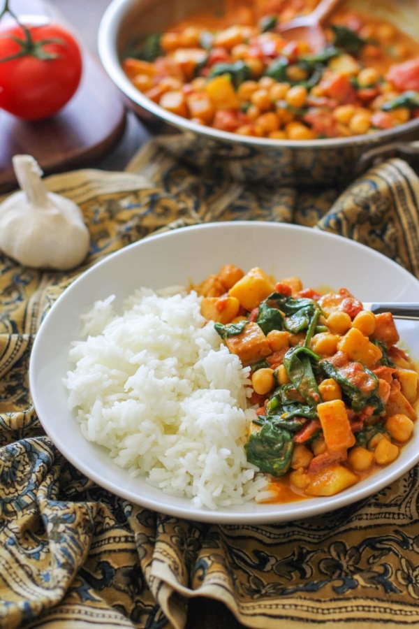 White bowl with chana saag and white rice inside. An ornate decorative napkin underneath the bowl and a skillet full of chickpea curry in the background.