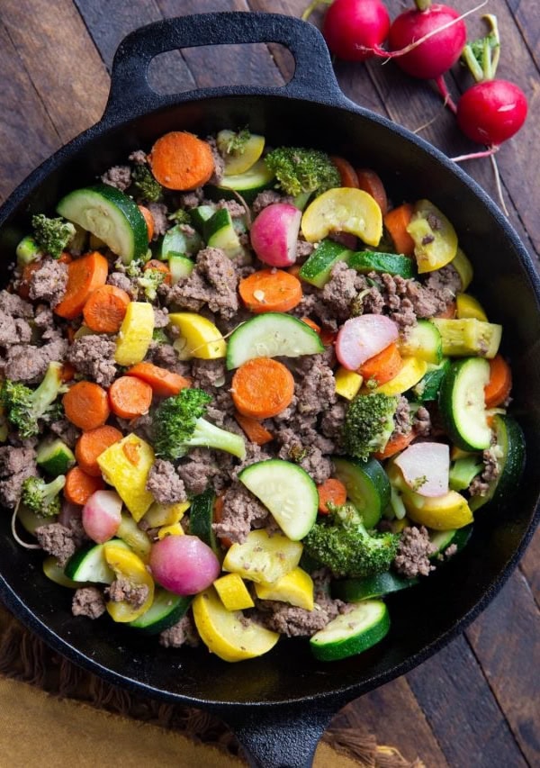 Close up of a cast iron skillet with ground beef and vegetables inside sitting on a wooden backdrop with a golden napkin.