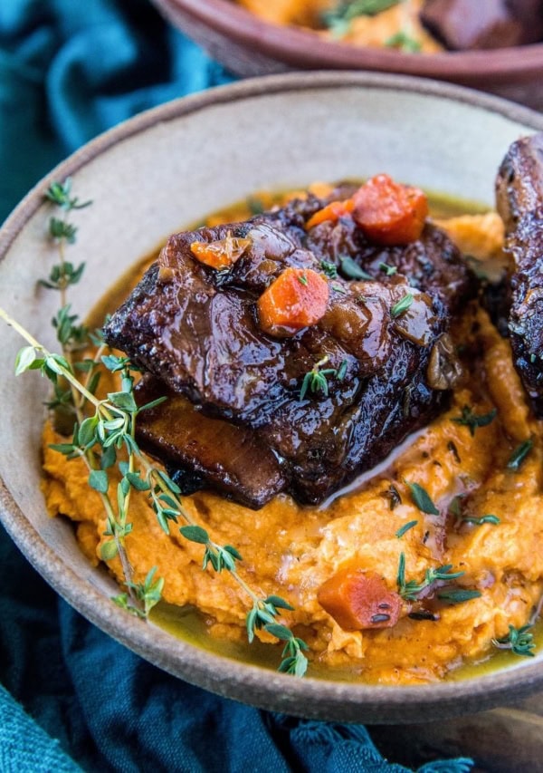 Beef short ribs on top of mashed sweet potatoes in a large bowl, ready to eat! A dark blue napkin to the side and a rustic wood backdrop.