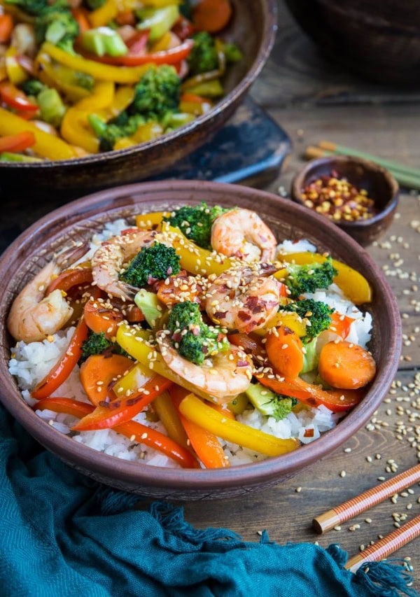 Big bowl of shrimp stir fry with a dark blue napkin to the side with chopsticks and a skillet of stir fry in the background.