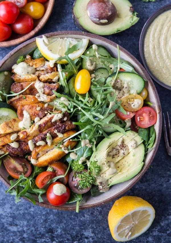 Top down image of whole bowl of crispy chicken salad with avocado, arugula, lemon tahini dressing, and a blue background.