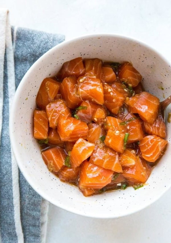 Bowl of salmon poke with spoon and blue napkin on white surface