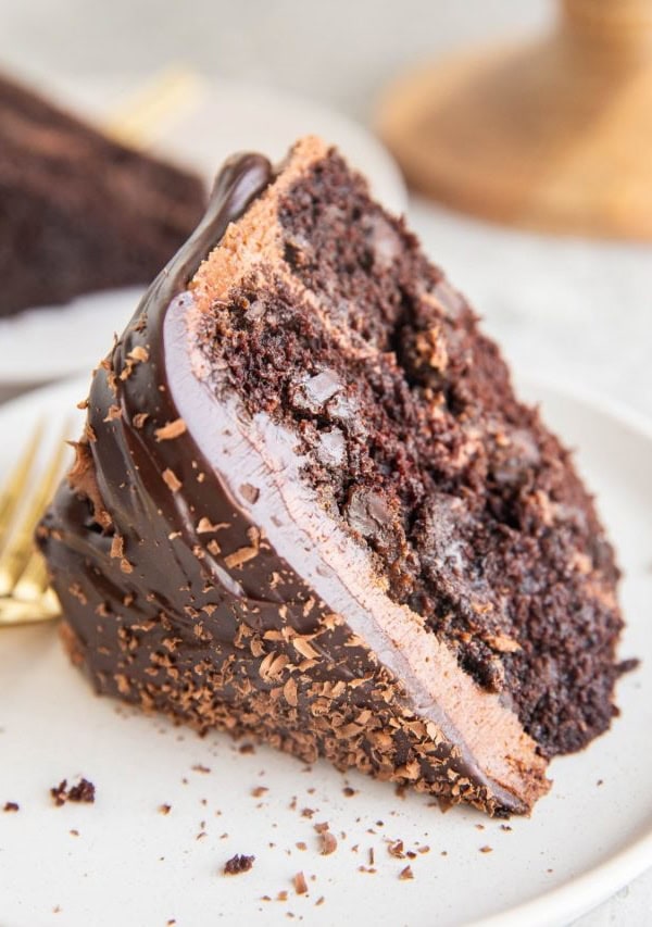 Thick slice of chocolate layer cake with chocolate frosting and chocolate ganache on a white plate with a gold fork.