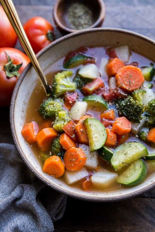 Big ceramic bowl of healthy vegetable soup with a gold spoon, ready to eat. A grey napkin and a fresh tomato to the side.