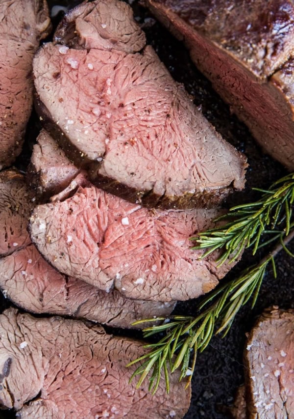 Close up top down photo of medium-rare beef tenderloin cut into slices laying in a cast iron skillet with fresh rosemary and sea salt sprinkled on top.