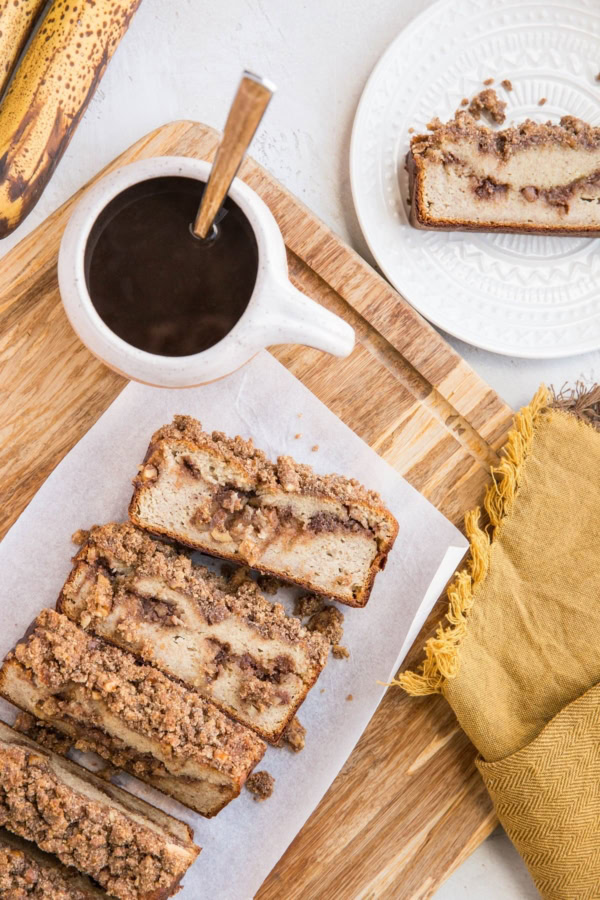 Grain-Free Cinnamon Swirl Banana Bread made paleo-friendly with almond flour and pure maple syrup. Sitting on a cutting board cut into slices with a white plate with a single slice, ready to eat. A gold napkin and cup of coffee to the side.