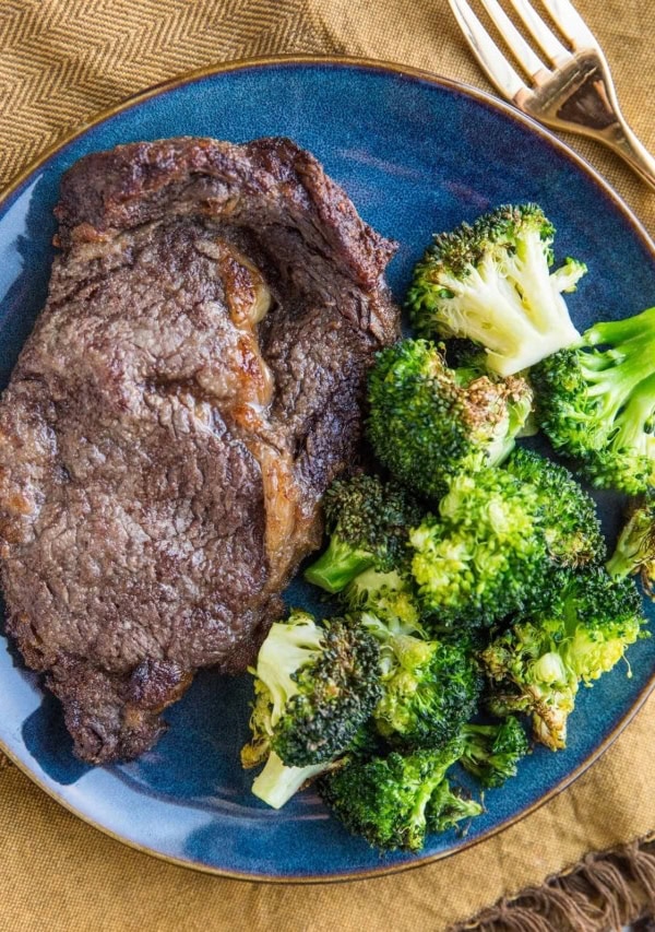 Blue plate with steak and broccoli on it with a gold fork to the side and a golden napkin