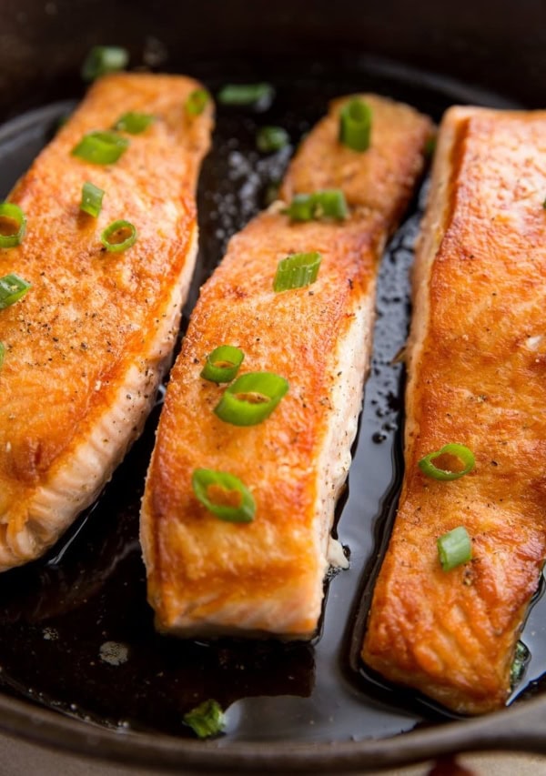 Three salmon filets in a cast iron skillet looking crispy and delicious with green onion sprinkled on top