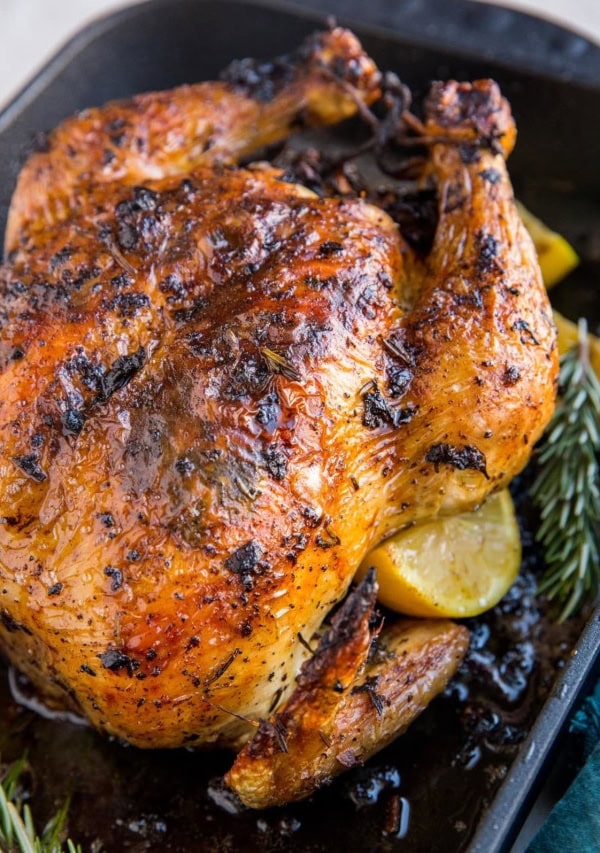 Roasting pan with roast chicken inside and lemon slices and rosemary with a dark blue napkin to the side