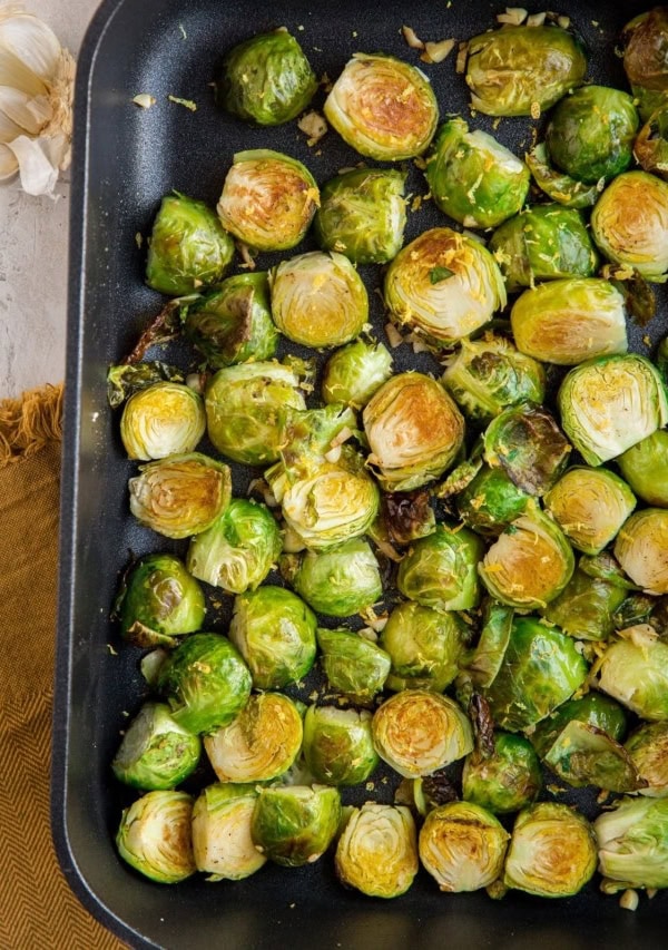 Roasted Brussel Sprouts in a cast iron casserole dish with lemon and garlic off to the side and a brown napkin