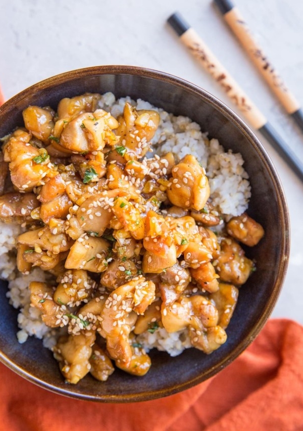 top down photograph of honey garlic chicken over brown rice in a blue bowl with a red napkin and chopsticks off to the side