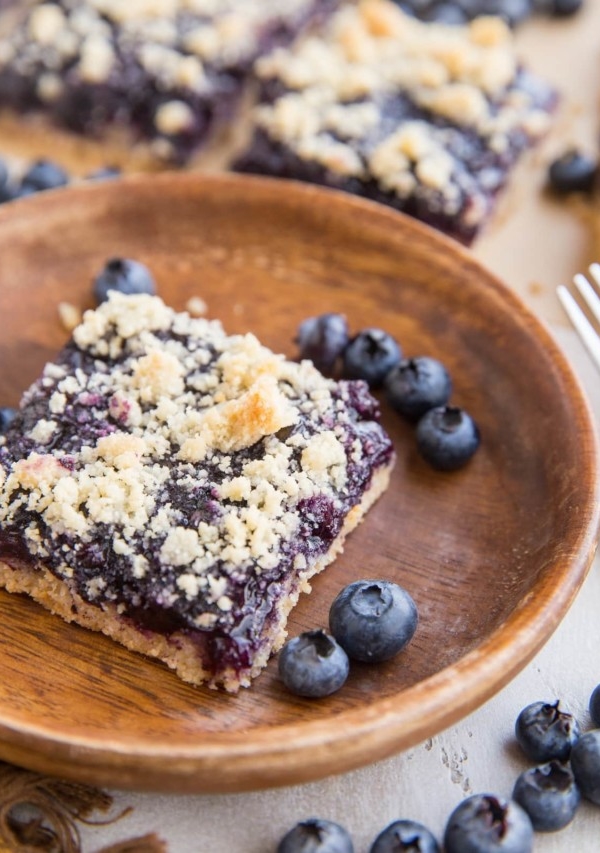 Keto blueberry crumb bars in the background, one bar on a wooden plate in the front with fresh blueberries all around