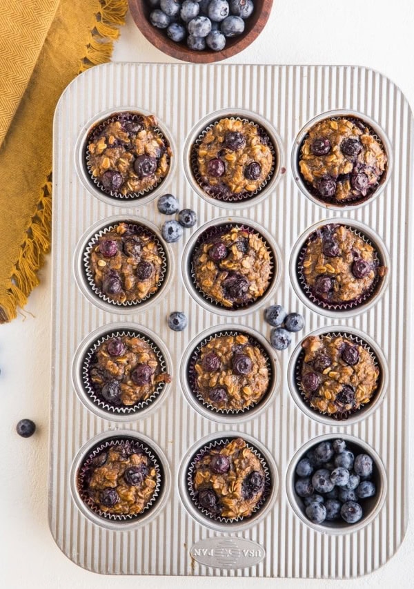 Blueberry Banana Baked Oatmeal Cups in a muffin tray with fresh blueberries to the side and a golden napkin