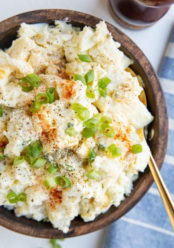 wooden bowl of potato salad with green onions on top and a spoon. Blue napkin off the the side and pepper grinder.