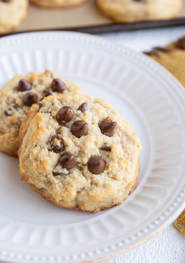 White plate with two coconut flour cookies on top with tray of cookies in the background.