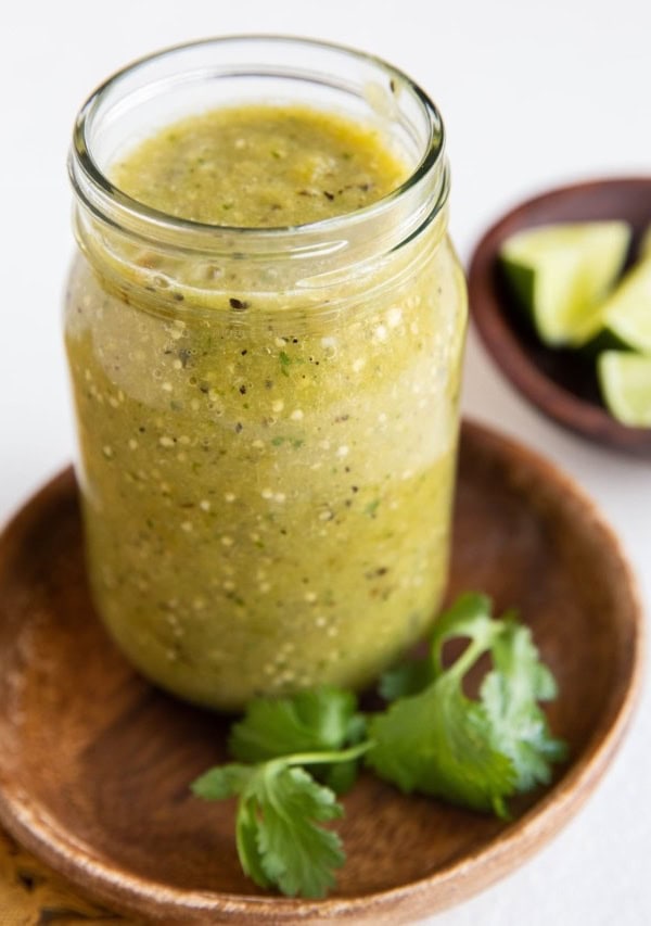 Jar of green tomatillo salsa on a wooden plate with bowl of limes in the background