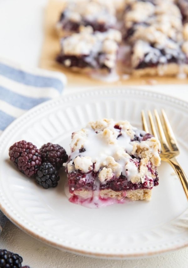 White plate with a blackberry crumb bar and fresh blackberries to the side and a gold fork.