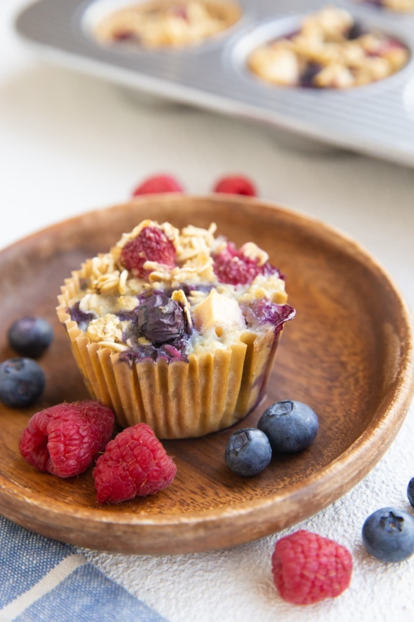 Berry baked oatmeal muffin on a wooden plate with fresh berries all around and more muffins in the background