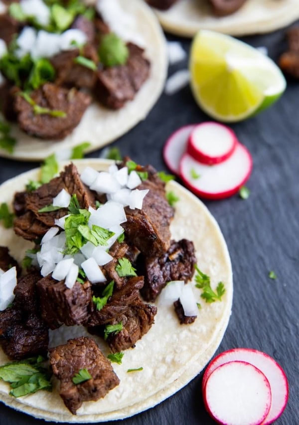 three carne asada tacos on a black slate with slices of turnips and limes to the sides