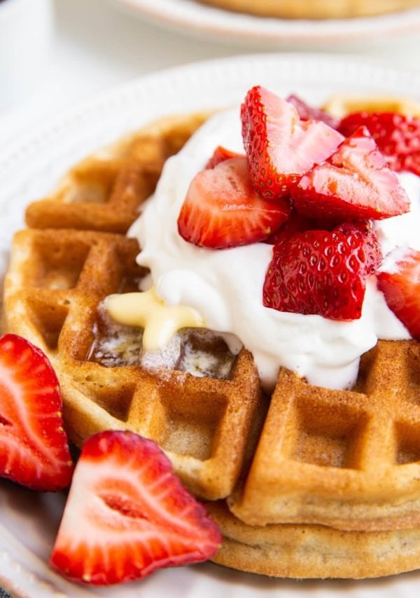 Stack of Belgian waffles on a white plate with whipped cream, butter, and strawberries