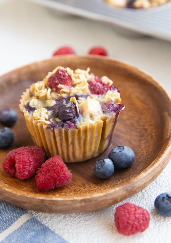 Berry baked oatmeal muffin on a wooden plate with fresh berries all around and more muffins in the background