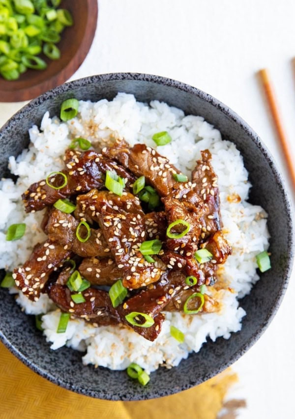 Black bowl with white rice and Korean beef sprinkled with sesame seeds and green onions. Chop sticks to the side, a bowl of green onions and a golden napkin