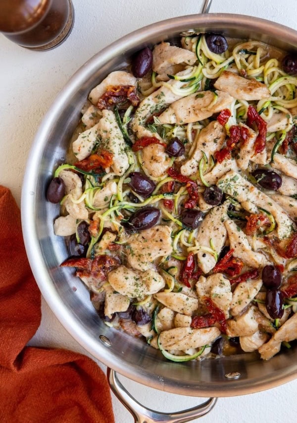 Stainless steel skillet with creamy sun-dried tomato chicken and zoodles with a red napkin to the side and a pepper shaker