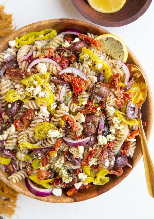 Wooden bowl of Greek pasta salad with a gold spoon, bowl of lemons and golden napkin