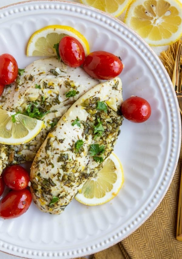 Two chicken breasts on a white plate with cherry tomatoes, sliced lemons and golden napkin