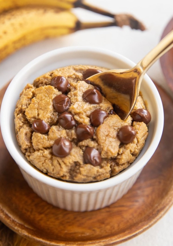 Ramekin of baked oats with chocolate chips on top and a golden spoon with ripe bananas in the background