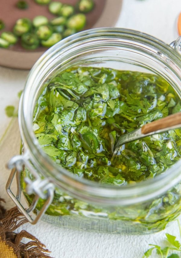 Jar of chimichurri sauce with a napkin to the side, along with fresh cilantro and jalapenos in the background.