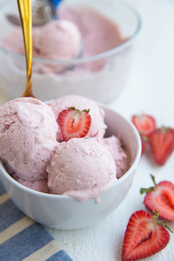 White bowl of strawberry ice cream with a gold spoon and fresh strawberries to the side.
