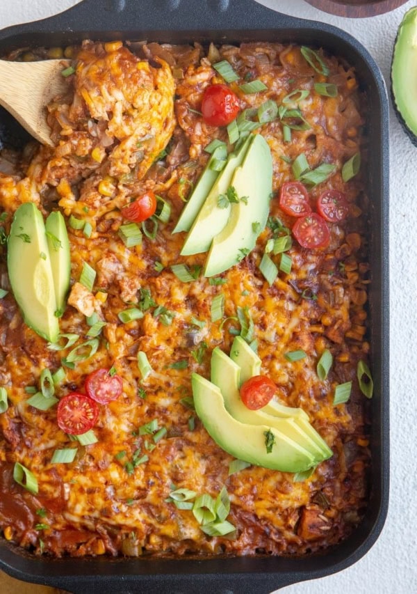 Top down image of cheesy enchilada casserole with a wooden spoon about to serve.