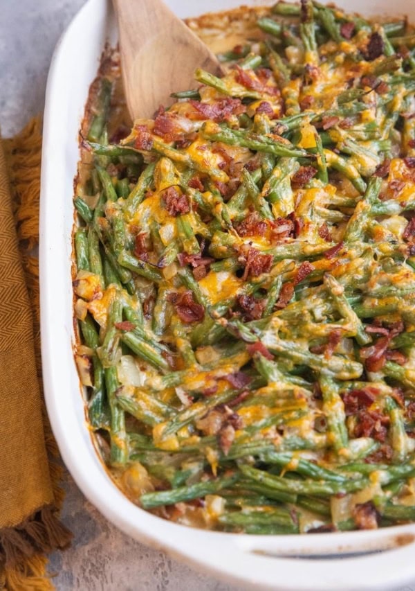 Large casserole dish of cheesy green bean casserole with chopped cooked bacon sprinkled on top and a napkin off to the side.