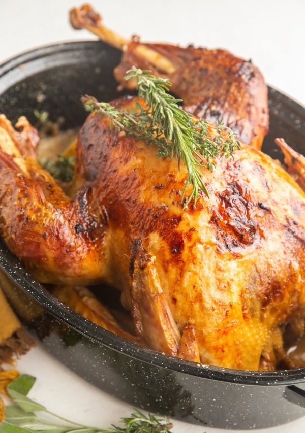 Roast turkey in a roasting pan with fresh herbs on top and fresh herbs to the side.