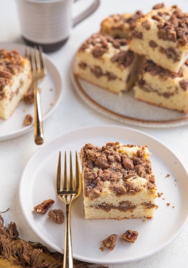 Two white plates with slices of coffee cake on top with a plate of several slices of coffee cake and a cup of coffee to the side.