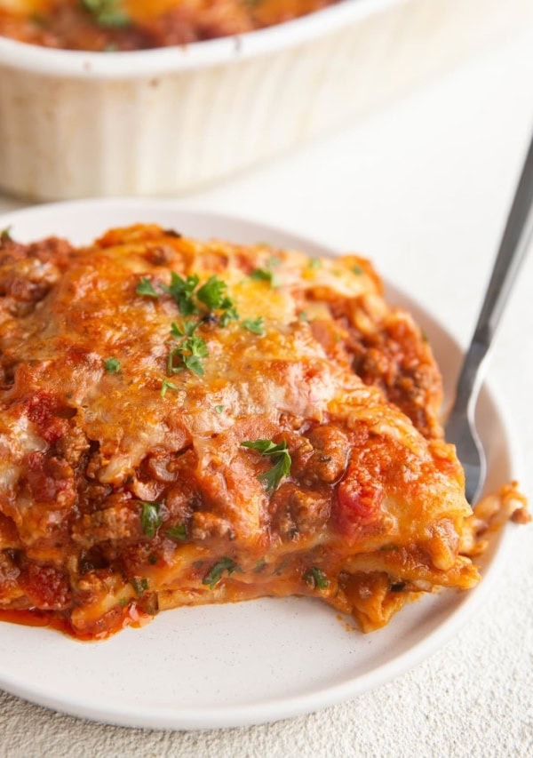 White plate of a large slice of gluten-free lasagna with the casserole dish of lasagna in the background.