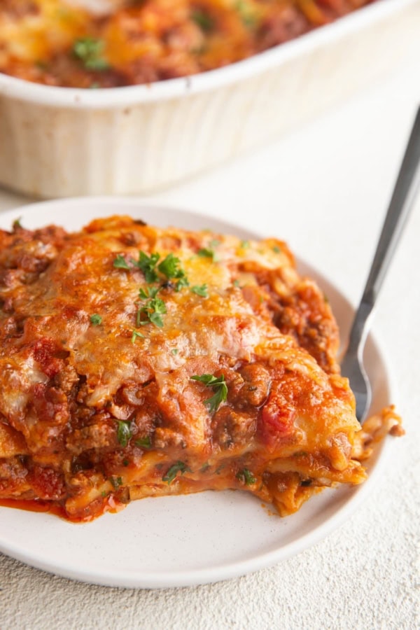 White plate of a large slice of gluten-free lasagna with the casserole dish of lasagna in the background.