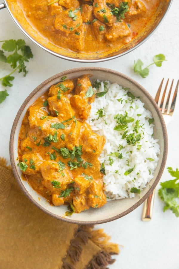 Bowl of chicken tikka masala with rice and a serving dish of chicken tikka masala with a gold fork and a gold napkin.