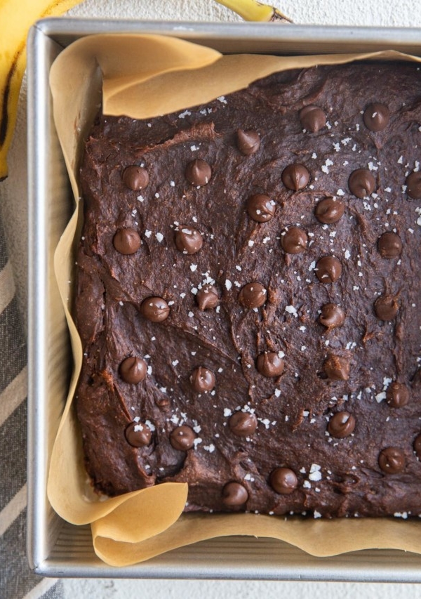 Square baking pan with healthy banana brownies sprinkled with sea salt and chocolate chips fresh out of the oven. A fresh banana and a napkin to the sides of the pan.