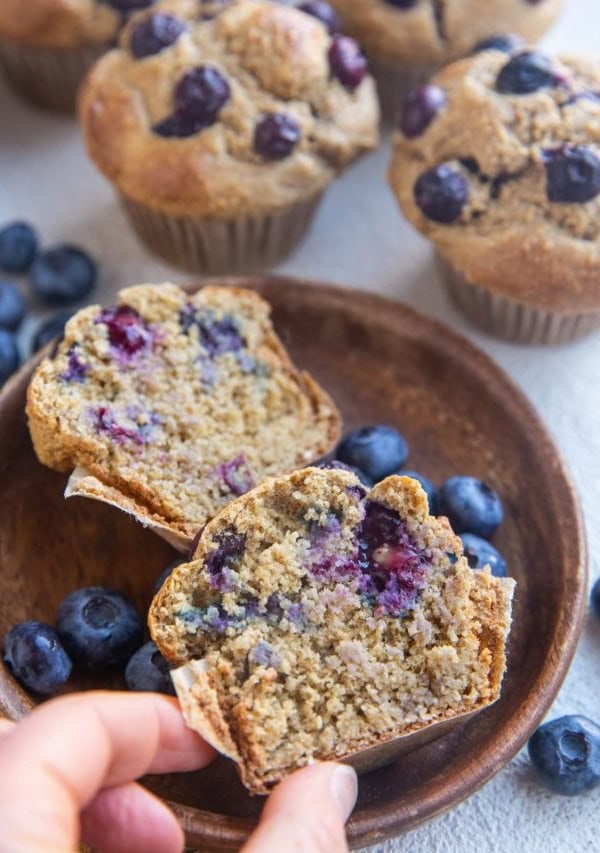 Plate with a muffin sliced in half and a hand picking up one of the halves, with full muffins in the background.