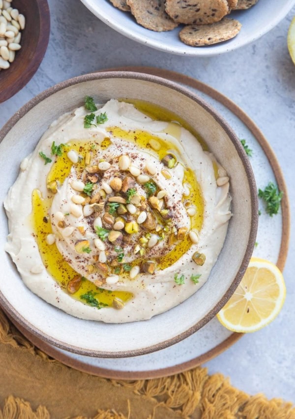 Bowl of hummus with pine nuts, pistachios and oil on top with a bowl of crackers to the side.