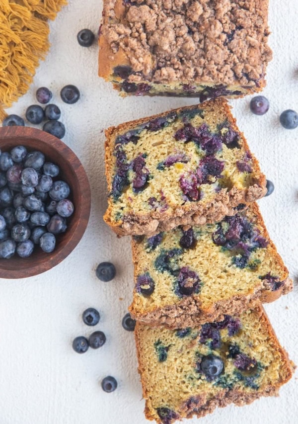 Cut up loaf of blueberry bread on a white background with fresh blueberries all around.