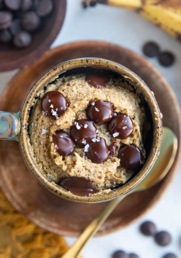 Mug full of banana mug cake sitting on a wooden plate with chocolate chips and ripe bananas to the side and a gold spoon.