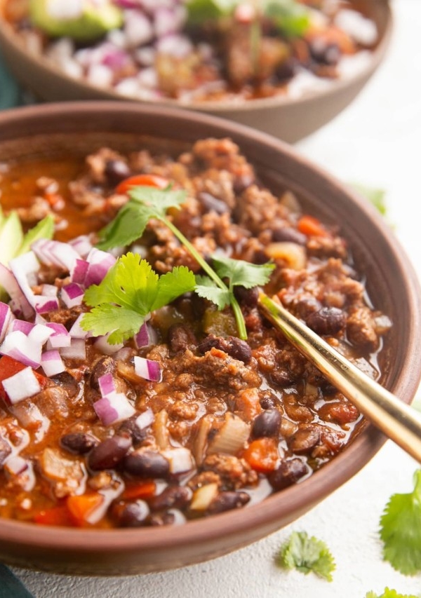 Two bowls of beef chili with a gold spoon, sprinkled with red onions and cilantro.